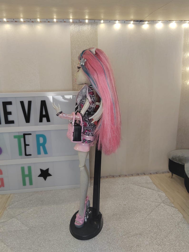 May include: A doll with long pink hair featuring blue streaks, wearing a patterned top and skirt, a small black and pink handbag, and pink platform shoes. The doll is positioned on a black stand in front of a light box displaying the text EVA TER H.