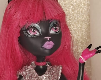 Può includere: Una bambola con pelle nera, orecchie da gatto a punta e lunghi capelli rosa acceso. La bambola ha occhi rosa, labbra viola chiaro e indossa un top argentato testurizzato con un girocollo rosa. Una mano è sollevata, mostrando unghie nere e un braccialetto.