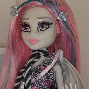 May include: A doll with light grey skin, pink and teal hair, and silver fleur-de-lis hair clips. The doll features pink lips, dark eye makeup, and a black and white striped necktie. It wears a patterned dress with pink accents and has visible wings.