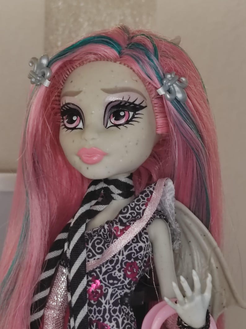 May include: A doll with light grey skin, pink and teal hair, and silver fleur-de-lis hair clips. The doll features pink lips, dark eye makeup, and a black and white striped necktie. It wears a patterned dress with pink accents and has visible wings.