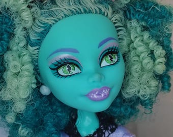 Peut inclure: Gros plan sur une poupée à la peau turquoise, aux yeux verts et aux cheveux bouclés turquoise et vert menthe. La poupée porte un haut lavande avec un col en dentelle noire et un petit chapeau rose. Les lèvres de la poupée sont violet clair.