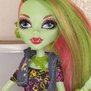 Puede incluir: Muñeca Monster High con piel verde, ojos azules y cabello rosa y verde brillante. Viste un vestido negro con un diseño rosa y amarillo neón, una chaqueta vaquera y accesorios verdes. Sus labios son rojos.