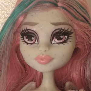 May include: A doll with pale grey skin, freckles, and pink lips. The doll has long, straight hair in shades of pink and teal. The eyes are large with pink irises, dark eyeliner, and long eyelashes. The doll features sculpted eyebrows and a small nose.