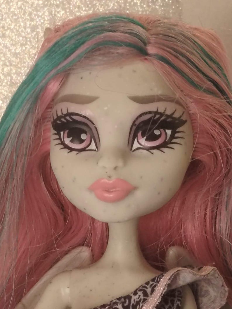May include: A doll with pale grey skin, freckles, and pink lips. The doll has long, straight hair in shades of pink and teal. The eyes are large with pink irises, dark eyeliner, and long eyelashes. The doll features sculpted eyebrows and a small nose.