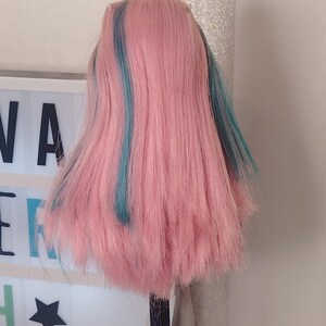 May include: A doll with long, straight pink hair featuring vibrant teal blue streaks. The hair is styled with a centre part and falls past the shoulders. The doll is positioned in front of a white light box displaying the partial text NA ER H.