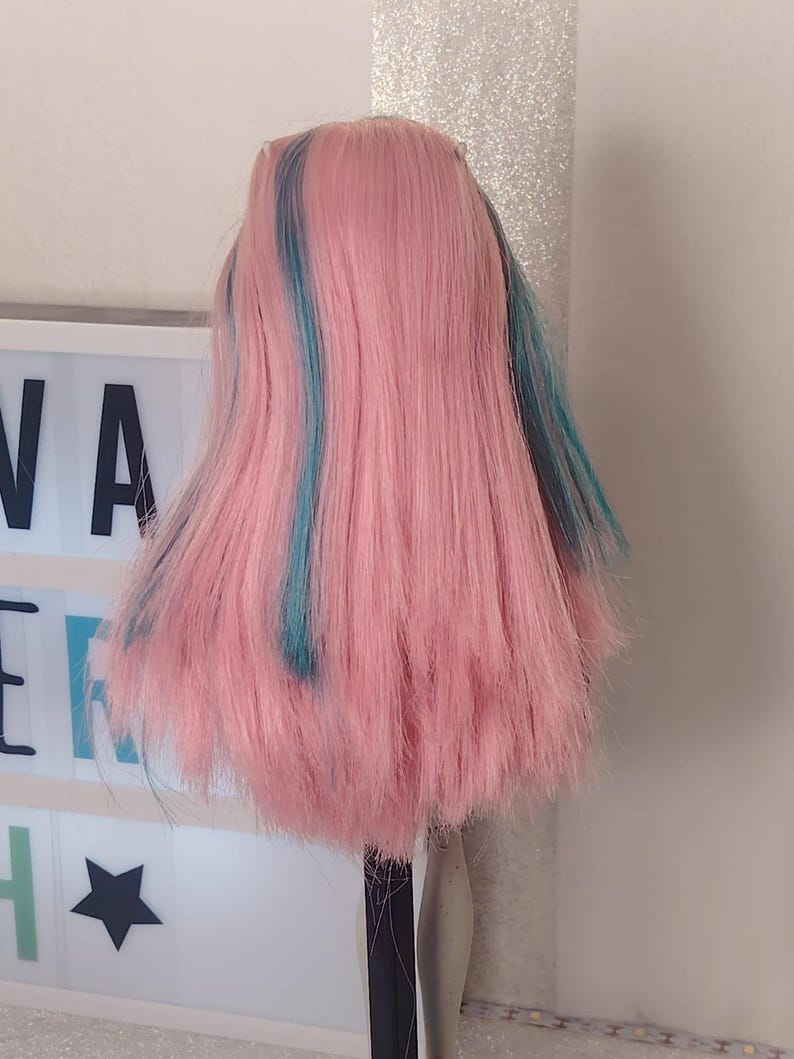 May include: A doll with long, straight pink hair featuring vibrant teal blue streaks. The hair is styled with a centre part and falls past the shoulders. The doll is positioned in front of a white light box displaying the partial text NA ER H.