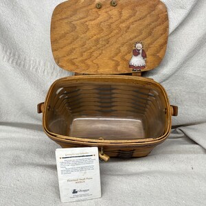 May include: A brown wicker basket with a wooden lid. The lid has a painted image of a woman in a red dress and white apron. The basket is labeled "Heartland Small Purse Basket".