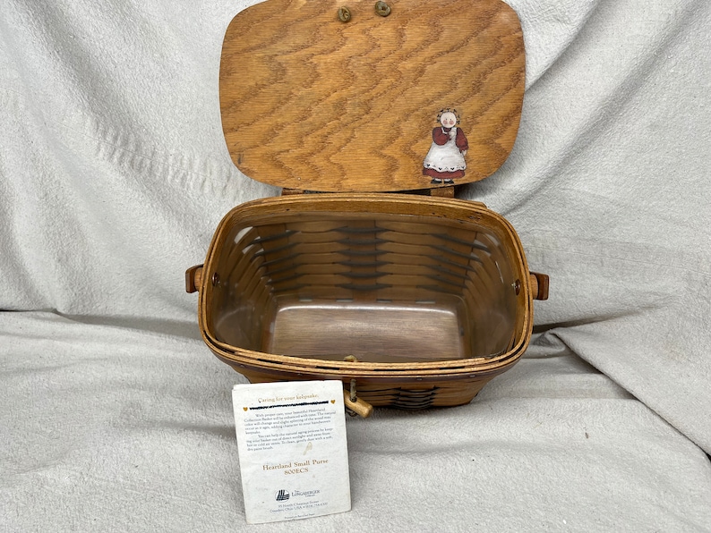 May include: A brown wicker basket with a wooden lid. The lid has a painted image of a woman in a red dress and white apron. The basket is labeled "Heartland Small Purse Basket".