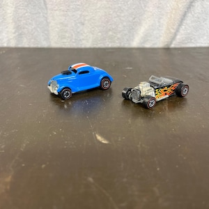 May include: Two toy cars, one blue with a white stripe and one black with flames. Both cars have black wheels.