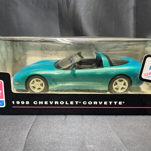 Puede incluir: Un coche modelo Chevrolet Corvette de 1998, de color turquesa, en una vitrina de pl&aacute;stico transparente. El coche tiene un interior negro y ruedas plateadas. La caja tiene el logotipo de Chevrolet y el texto "1998 Chevrolet Corvette".