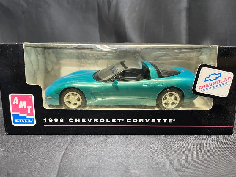 Puede incluir: Un coche modelo Chevrolet Corvette de 1998, de color turquesa, en una vitrina de pl&aacute;stico transparente. El coche tiene un interior negro y ruedas plateadas. La caja tiene el logotipo de Chevrolet y el texto "1998 Chevrolet Corvette".