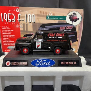 May include: A black 1953 Ford F-100 panel van model with red wheels, labeled "Fire Chief Gasoline." The model is on a black display stand with the Ford logo and "Precision Model" text. The box behind it reads "Limited Edition 1953 F-100 Delivery Van."