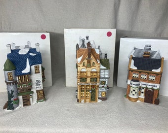May include: Three ceramic miniature buildings with snow-covered roofs. One has a blue roof and reads "The Wood Shop Yarn". Another reads "Counting House" and the third reads "No. Heeton Watchmaker".