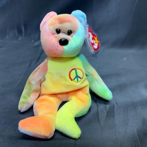 May include: A colorful plush bear toy with a peace sign on its chest. The bear features a rainbow of colors, including pink, orange, yellow, green, and blue. It has a black nose and eyes, and a Ty tag attached to its ear.