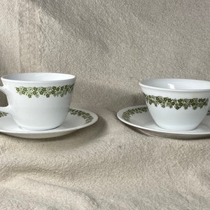 Peut inclure: Deux tasses à thé et soucoupes blanches avec un motif floral vert. Les tasses ont une forme large et arrondie et une poignée sur le côté droit. Les soucoupes ont un motif floral assorti.