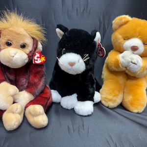 Puede incluir: Tres peluches: un mono con un atuendo a cuadros rojos, un gato blanco y negro y un oso amarillo dorado con las patas juntas. Cada juguete tiene una etiqueta.