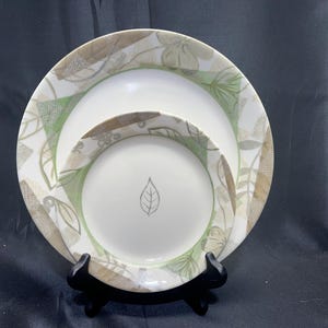 May include: Two white ceramic plates with a leaf design. The plates are decorated with a green and tan leaf pattern around the rims. The smaller plate has a leaf illustration in the center. The plates are displayed on a black stand.