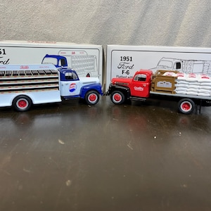 May include: Two vintage 1951 Ford F-6 trucks, one white with blue accents and a Pepsi Cola logo, the other red with a brown wooden bed and white bags of flour. Both trucks are diecast models.