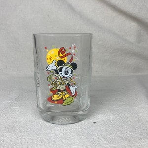May include: A clear glass with a Mickey Mouse design. Mickey is wearing a safari outfit and holding a pair of binoculars. The glass has a red, yellow, and green design with confetti and the text "2000" at the bottom. The glass is made by Disney.