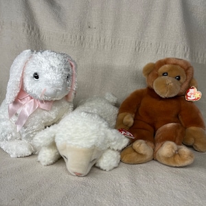 May include: Three plush toys: a white bunny with pink ears and a pink ribbon, a white lamb with its head resting on the ground, and a brown monkey with a red heart tag.