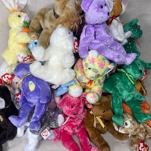 Puede incluir: Una colección de peluches Beanie Babies de colores en varias formas de animales, incluyendo un conejito, un oso, un perro y un gato. Los Beanie Babies están dispuestos en una pila, con algunos de ellos asomando por detrás de otros.