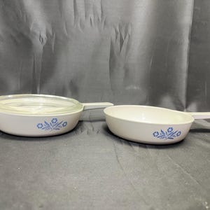 May include: Two white casserole dishes with blue floral designs. One dish has a clear glass lid. Both dishes have a handle on one side. The dishes are likely made of ceramic or glass.