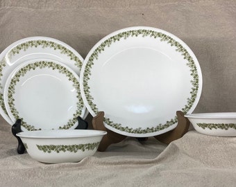 May include: Set of white ceramic dinnerware with a green floral pattern. The set includes a large dinner plate, a salad plate, a bowl, and a smaller bowl.