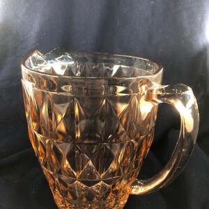 May include: A pink glass pitcher with a diamond pattern. The pitcher has a handle and a spout.