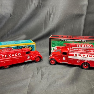 May include: Two red Texaco tanker trucks, one with a streamlined design and the other a 1939 Studebaker model, are displayed. The trucks have the Texaco logo and are accompanied by their original boxes.