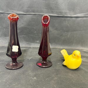 Puede incluir: Dos floreros de vidrio rojo oscuro, altos y delgados, con bordes acampanados y ondulados y bases circulares, junto a una pequeña figura de pájaro de vidrio amarillo brillante con un acabado liso y mate, con cola y pico detallados.