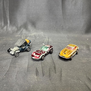 May include: Three vintage toy cars are displayed. A dark blue hot rod with an open top, a red and white sports car, and a gold-colored sports car with a red interior. The cars are on a dark surface.