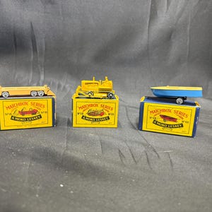 May include: Three vintage Matchbox Series toy vehicles on their original boxes. The toys include a yellow flatbed truck, a yellow bulldozer, and a blue and yellow boat. The boxes are yellow with blue accents and feature the text "MATCHBOX SERIES" and "A MOKO LESNEY."
