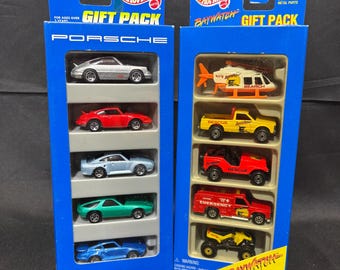 Puede incluir: Dos paquetes de regalo Hot Wheels en cajas de cartón azul. El paquete izquierdo contiene cinco coches Porsche a escala en plata, rojo, azul claro, verde y azul. El paquete derecho incluye vehículos de rescate con temática Baywatch.