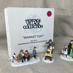 May include: A set of three hand-painted porcelain figurines depicting a market scene. The figurines are part of the Heritage Village Collection and are titled "Market Day".