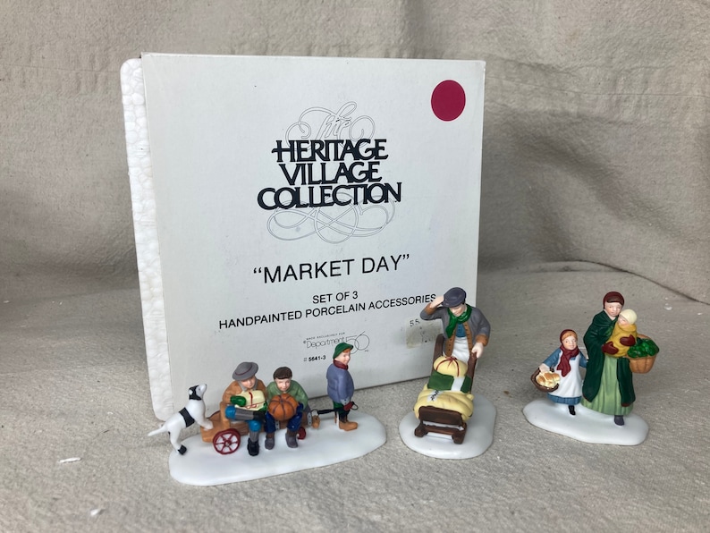May include: A set of three hand-painted porcelain figurines depicting a market scene. The figurines are part of the Heritage Village Collection and are titled "Market Day".