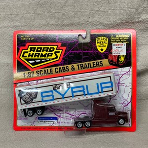 May include: A 1:87 scale model of a semi-truck and trailer with a Hershey's Syrup advertisement. The truck is maroon and the trailer is white with blue and red accents. The packaging is red and white with the Road Champs logo.
