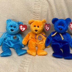 May include: Three Ty Beanie Baby plush toys. The first is blue with a swirl pattern and a gold coin on its chest. The second is orange with a purple ribbon and a gold crown on its chest. The third is blue with a gold ribbon and a gold star on its chest.