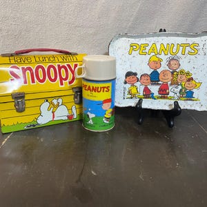 Peut inclure: Une boîte à lunch vintage Peanuts avec un design jaune et rouge représentant Snoopy. La boîte à lunch a une poignée et un loquet. Une gourde Peanuts avec un design bleu et blanc représentant Charlie Brown et Snoopy est également incluse. Un plateau en métal blanc avec un design Peanuts représentant les personnages de la bande dessinée est également inclus.