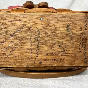 May include: A wooden Longaberger basket with a pink and white fabric lining. The bottom of the basket is signed with the text "Longaberger Baskets&reg; Handmade in Ohio USA".