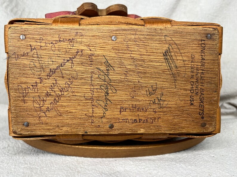 May include: A wooden Longaberger basket with a pink and white fabric lining. The bottom of the basket is signed with the text "Longaberger Baskets&reg; Handmade in Ohio USA".