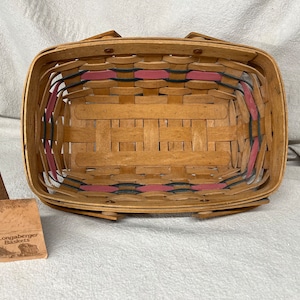 May include: A Longaberger Baskets brand woven basket with a natural wood finish and pink and black trim. The basket is empty and has a rectangular shape.