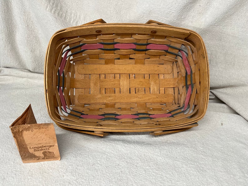 May include: A Longaberger Baskets brand woven basket with a natural wood finish and pink and black trim. The basket is empty and has a rectangular shape.