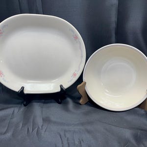 以下が含まれることがあります： 白の楕円形サービングプラッターと丸いボウル。どちらも縁に沿ってピンクと青の繊細な花柄があります。プラッターは黒いスタンドで支えられ、ボウルは木製のスタンドに置かれています。