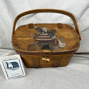 May include: A wooden Longaberger basket with a red, white, and blue painted design of a girl holding a flag. The basket has a wooden lid with a handle and a woven base. The tag reads "A Salute to Pride Longaberger Baskets The Longaberger Heartland Collection."