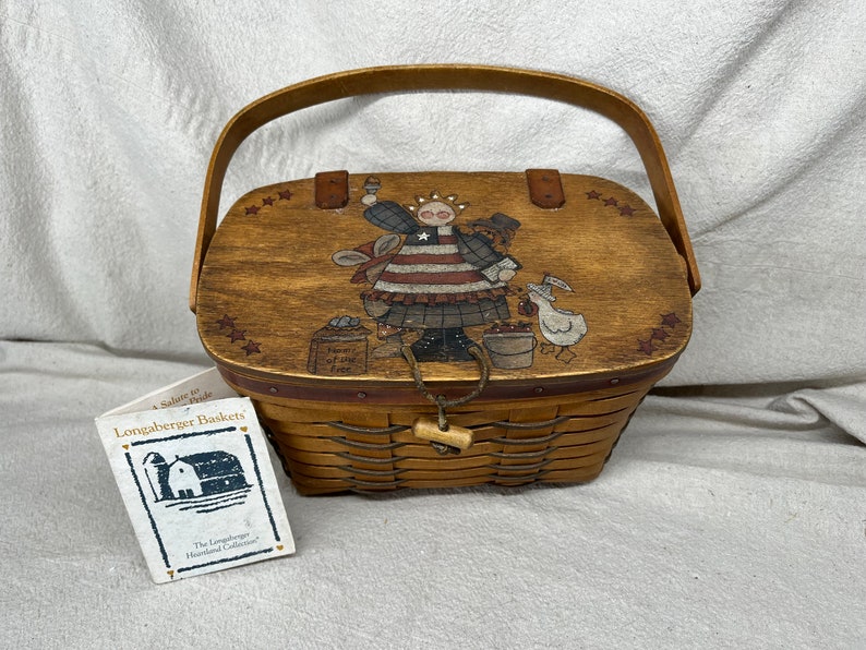 May include: A wooden Longaberger basket with a red, white, and blue painted design of a girl holding a flag. The basket has a wooden lid with a handle and a woven base. The tag reads "A Salute to Pride Longaberger Baskets The Longaberger Heartland Collection."