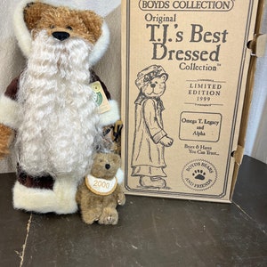 May include: A brown teddy bear wearing a white fur-trimmed coat and a white beard. The bear is holding a smaller brown teddy bear with a gold tag that says "2000". The bears are in front of a box that says "The Boyds Collection Original T.J.'s Best Dressed Collection Limited Edition 1999 Omega T. Legacy and Alpha Bears & Hares You Can Trust... Boyds Bears and Friends"