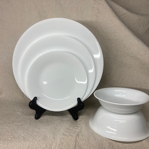 May include: A set of three white ceramic bowls, nested together, with a larger white ceramic bowl on a black stand.