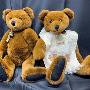 Puede incluir: Dos osos de peluche marrones, uno con una toalla blanca con un pequeño diseño bordado. El oso de la izquierda tiene una campana alrededor del cuello.