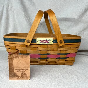 May include: A Longaberger basket with a wooden handle and a blue and pink woven design. The basket has a tag that reads "Remember Together 1993 Bee Basket".