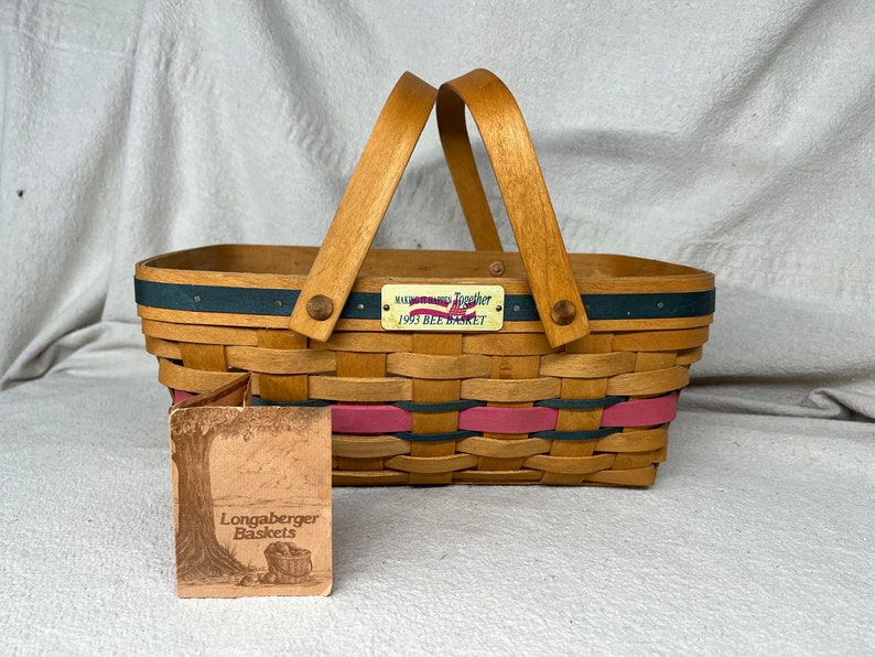 May include: A Longaberger basket with a wooden handle and a blue and pink woven design. The basket has a tag that reads "Remember Together 1993 Bee Basket".